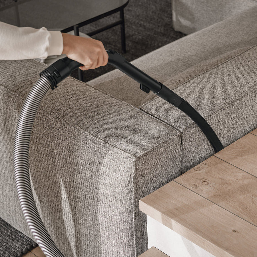 Person cleaning a gray sofa with a vacuum cleaner on a wooden floor.